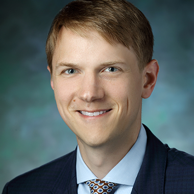 Kelly Mills, in a formal portrait, wearing a dark blue suit, light blue button down shirt and blue and white dotted tie